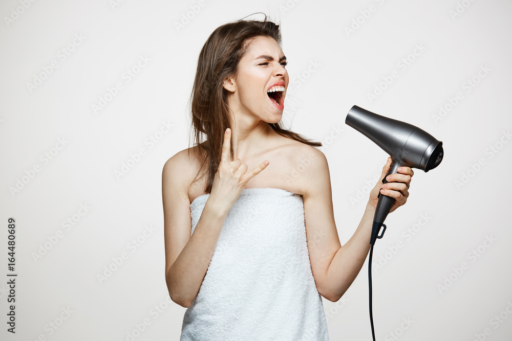 Cheerful beautiful girl in towel smiling laughing singing with hair dryer making funny face over white background. Beauty spa and cosmetology. Cheerful beautiful girl in towel smiling laughing singing with hair dryer making funny face over white background. Beauty spa and cosmetology.