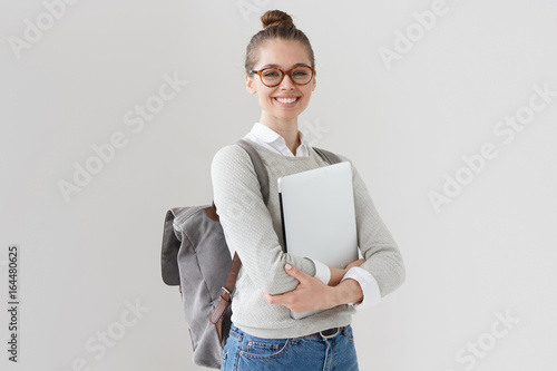 Indoor photo of college student girl isolated on gray background, smiling at camera, pressing laptop to chest, wearing backpack, ready to go to studies, start new project and suggest new ideas. photo