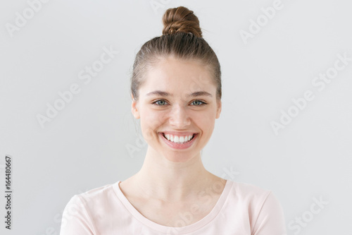 Indoor portrait of young attractive green-eyed female without makeup isolated on grey background in daylight smiling widely and happily, looking open and satisfied with communication and life.