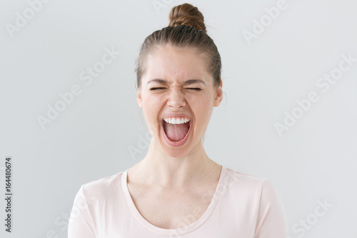 Indoor photo of young beautiful woman isolated on grey background screaming loudly with emotional expression on face with eyes closed and mouth open wide  looking deeply astonished with some news.