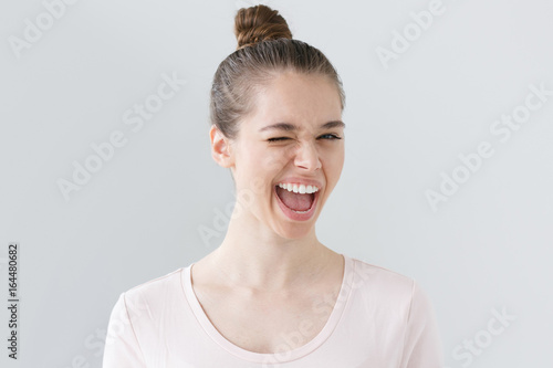 Indoor shot of attractive teenage girl isolated on grey background, looking happy and energetic, smiling with open mouth and winking as if she suggests great ideas for leisure time and activities.