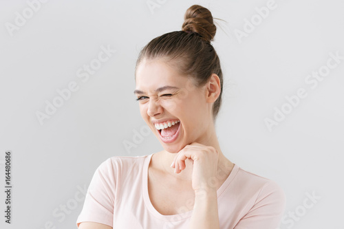 Tableau sur toile Closeup shot of beautiful teenage girl isolated on grey background, looking caught by hilarious joke, laughing with open mouth, winking, pressing chin with fingers, showing excitement and happiness