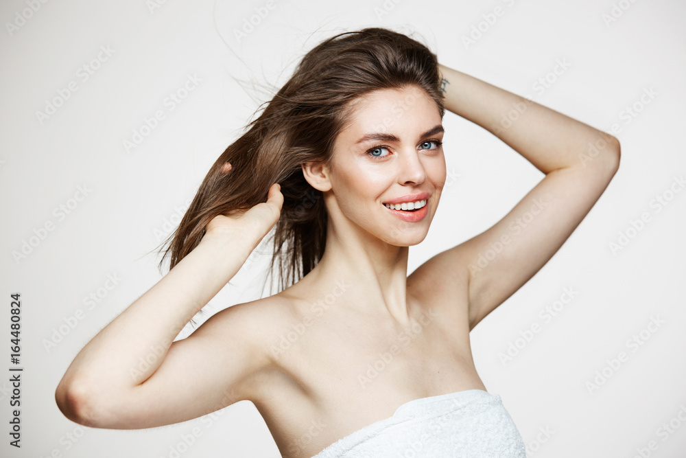 Beautiful young girl with perfect clean skin smiling looking at camera touching hair over white wall. Facial treatment.