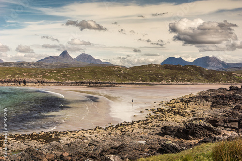 Assynt Peninsula, Scotland - June 7, 2012: Achnahaird Beach is sandy with gentle Atlantic Ocean waves out of Enard Bay. Lone walker far on beach. Mountainous background. Rocky foreground. Heavy clouds