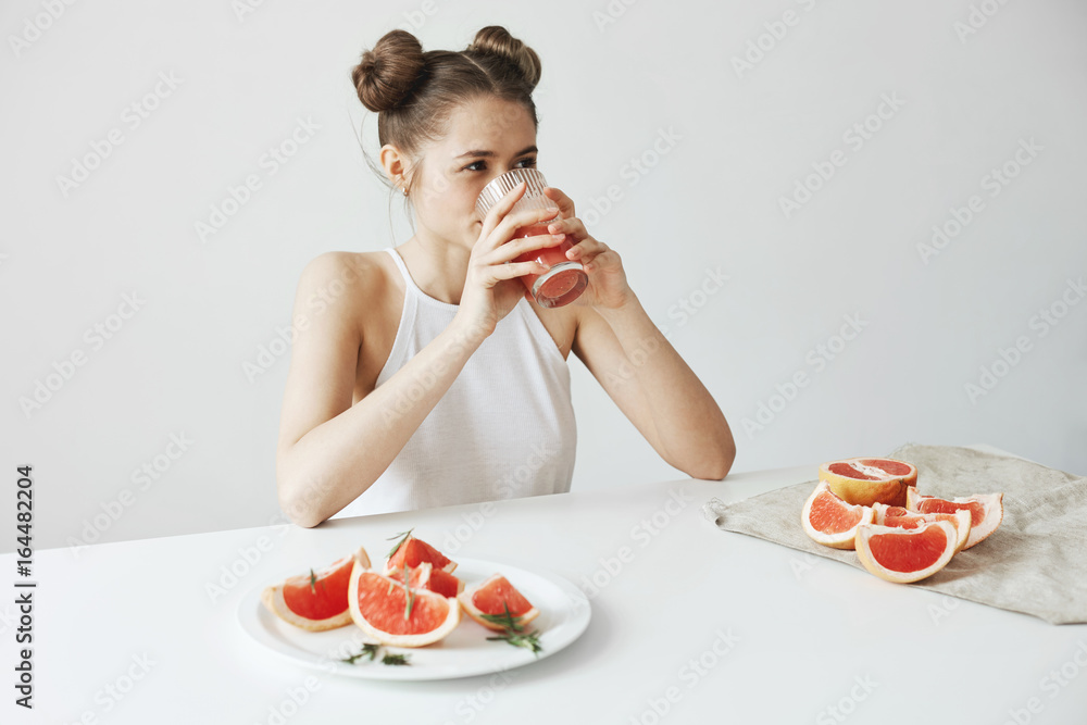 Happy beautiful girl smiling sitting at table drinking healthy detox fresh grapefruit smoothie over white wall. Happy beautiful girl smiling sitting at table drinking healthy detox fresh grapefruit smoothie over white wall.