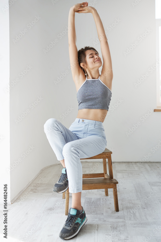 Young pretty girl with closed eyes smiling stretching sitting over white wall in morning. Young pretty girl with closed eyes smiling stretching sitting over white wall in morning.