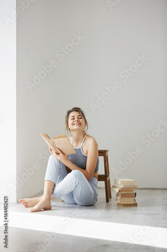 Young beautiful tender girl smiling holding book sitting on floor over white wall early in morning. photo