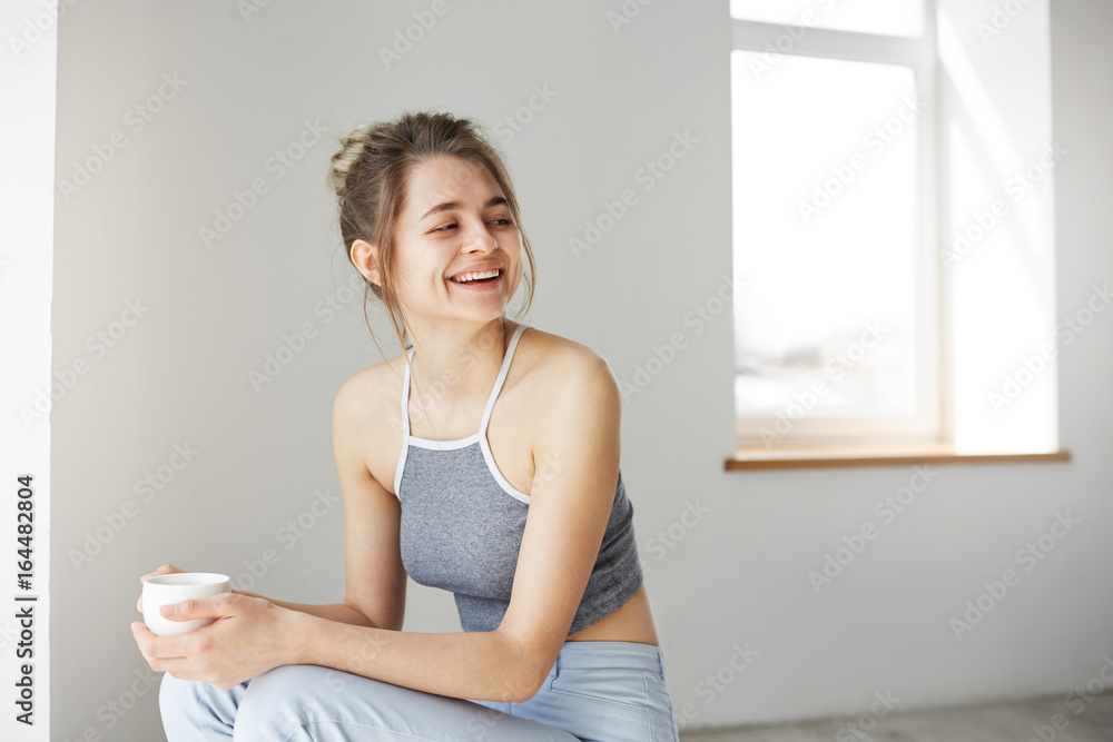 Portrait of young cheerful happy girl smiling laughing holding cup sitting on chair over white wall at home. Portrait of young cheerful happy girl smiling laughing holding cup sitting on chair over white wall at home.