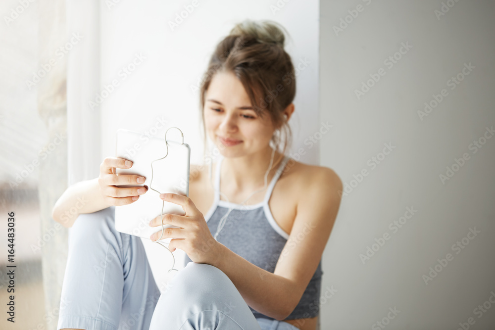 Portrait of young teenage cheerful girl in headphones smiling looking at tablet surfing web browsing internet sitting near window over white wall. Portrait of young teenage cheerful girl in headphones smiling looking at tablet surfing web browsing internet sitting near window over white wall.