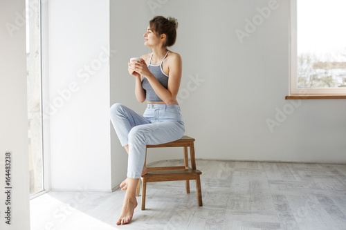 Young tender brunette girl smiling holding cup looking at window enjoying view sitting on chair over white wall in morning.
