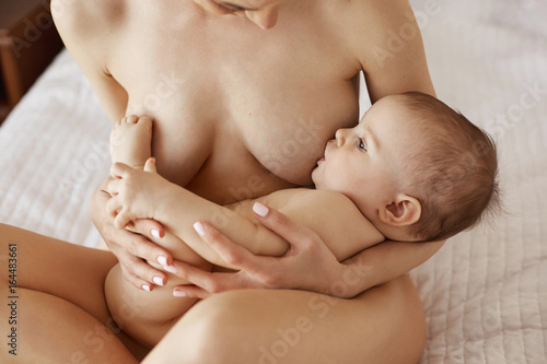 Young beautiful naked mom breastfeeding hugging her newborn baby smiling sitting on bed at home.