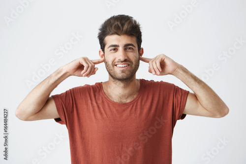 Portrait of annoyed young male Caucasian with eyes closed plugging his ears with fingers can't stand loud noise or ignoring stressful unpleasant situation or conflict. Negative human emotions.