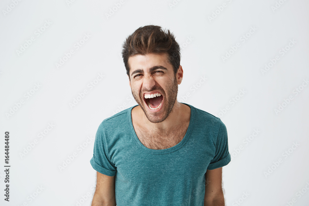 Indoor shot of furious young good-looking European man in casual cloth screaming with anger and rage, keeping his eyes closed. Student having an argument with his girlfriend. Human face expressions Indoor shot of furious young good-looking European man in casual cloth screaming with anger and rage, keeping his eyes closed. Student having an argument with his girlfriend. Human face expressions