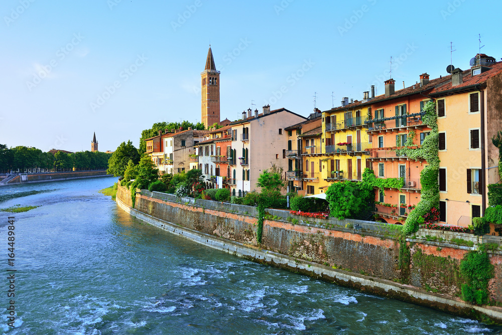 Beautiful facades of old houses on waterfront of the Adige River and bell tower of Santa Anastasia church in Verona Italy in morning sun. Verona is a popular tourist destination of Europe.