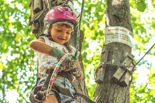 Happy 5 year old girl in pink protective helmet and equipment in a rope park in summer. © Masarik