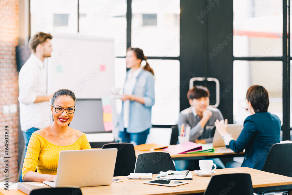 Smiling Asian woman using laptop computer, with young multiethnic business colleagues working together in background. Modern office job, teamwork brainstorm, or student team meeting concept