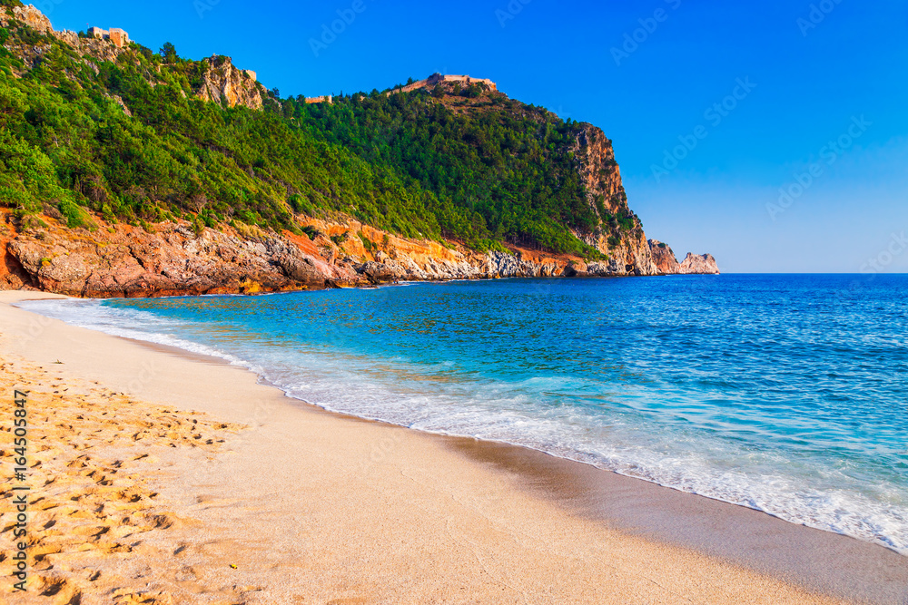 Cleopatra beach on sea coast with green rocks in Alanya peninsula, Antalya district, Turkey. Beautiful sunny landscape for tourism with clear water and sand. Alanya Castle on the cliff