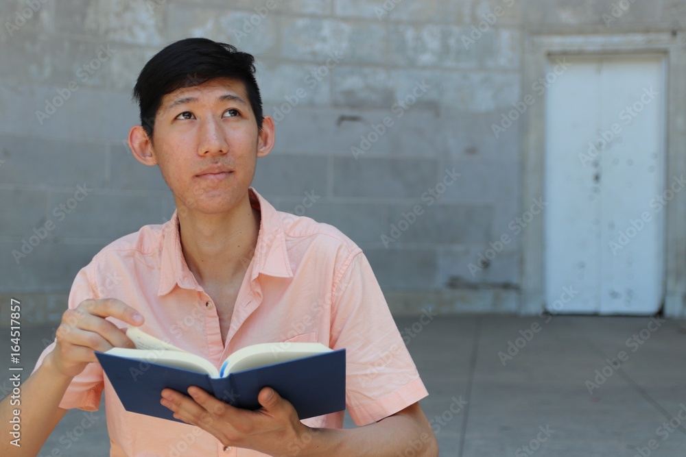 Young asian student reading a blue notebook while standing in front of gray concrete wall Young asian student reading a blue notebook while standing in front of gray concrete wall