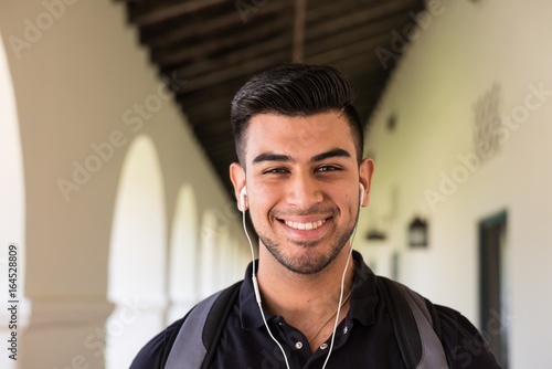 Slika na platnu Attractive male college student with a big smile on campus, wearing earphones an
