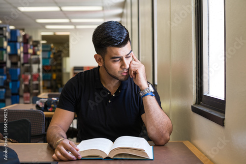 Canvas Print Tired male college student sitting in the library, reading a book, looking tired