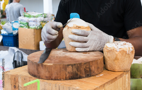 Hands with gloves trimming coconut flesh with long knife on wooden cutting board