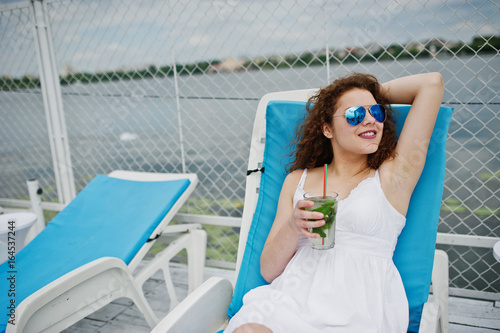 Portrait of an amazing young girl wearing sunglasses enjoying her cocktail sitting on a lounger in lakeside.