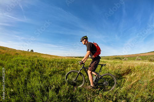 Young man riding on a bicycle on green meadow with a backpack © Aleksey