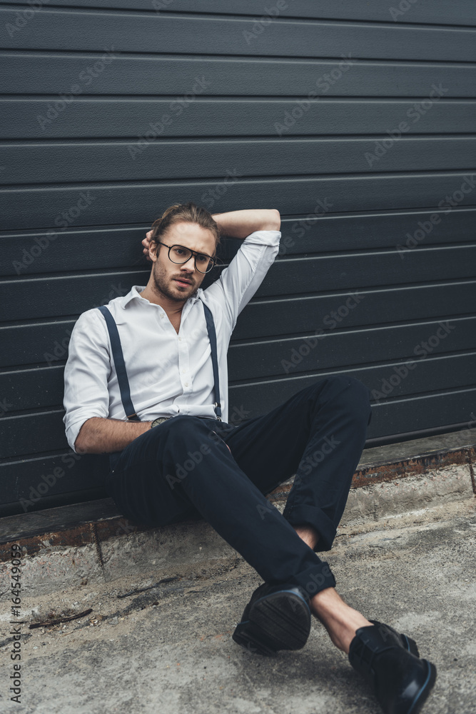 Handsome pensive stylish man in eyeglasses sitting with hand behind head on pavement and looking away Handsome pensive stylish man in eyeglasses sitting with hand behind head on pavement and looking away