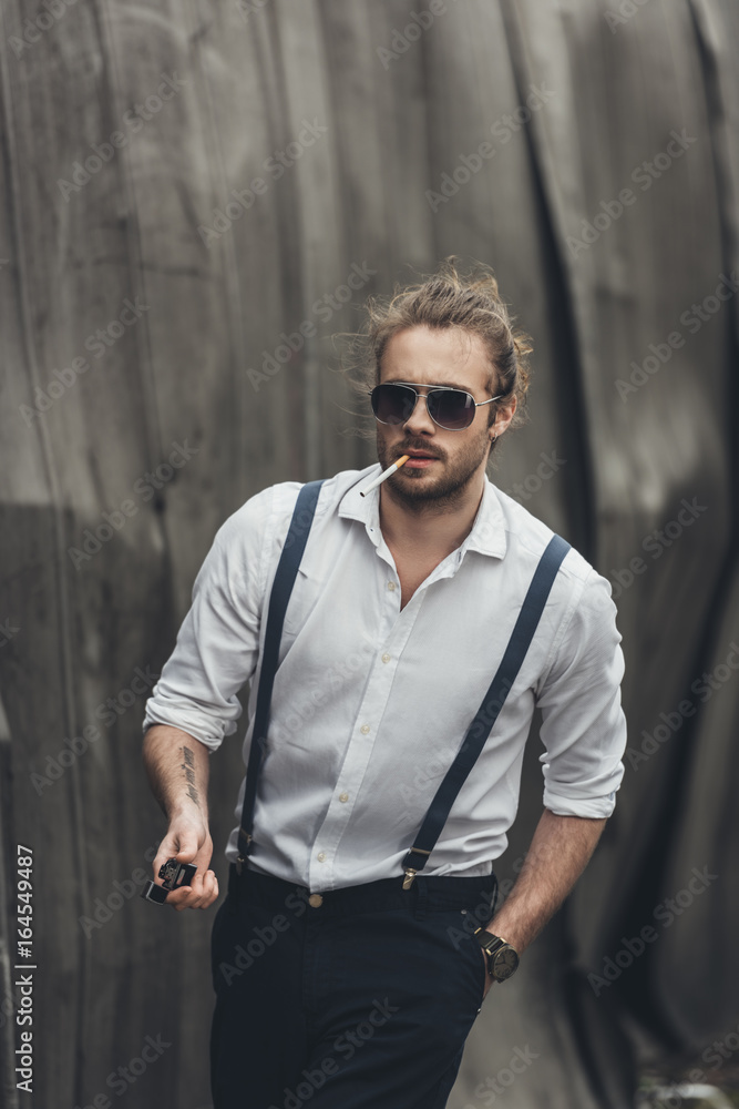 Obraz premium Stylish young man smoking cigarette and holding lighter while walking with hand in pocket