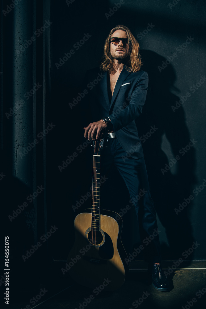 Handsome long haired man in sunglasses and stylish suit posing with acoustic guitar