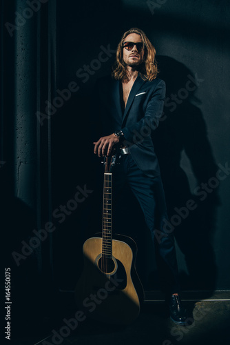 Handsome long haired man in sunglasses and stylish suit posing with acoustic guitar