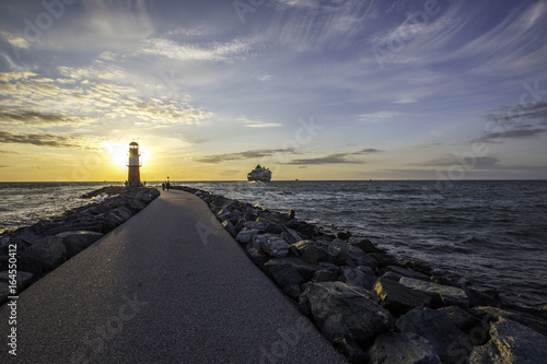 Schiff verl  sst den Hafen von Warnem  nde hinter einem Leuchtturm im Sonnenuntergang