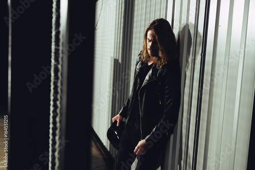 Pensive young long haired man holding helmet and leaning at wall