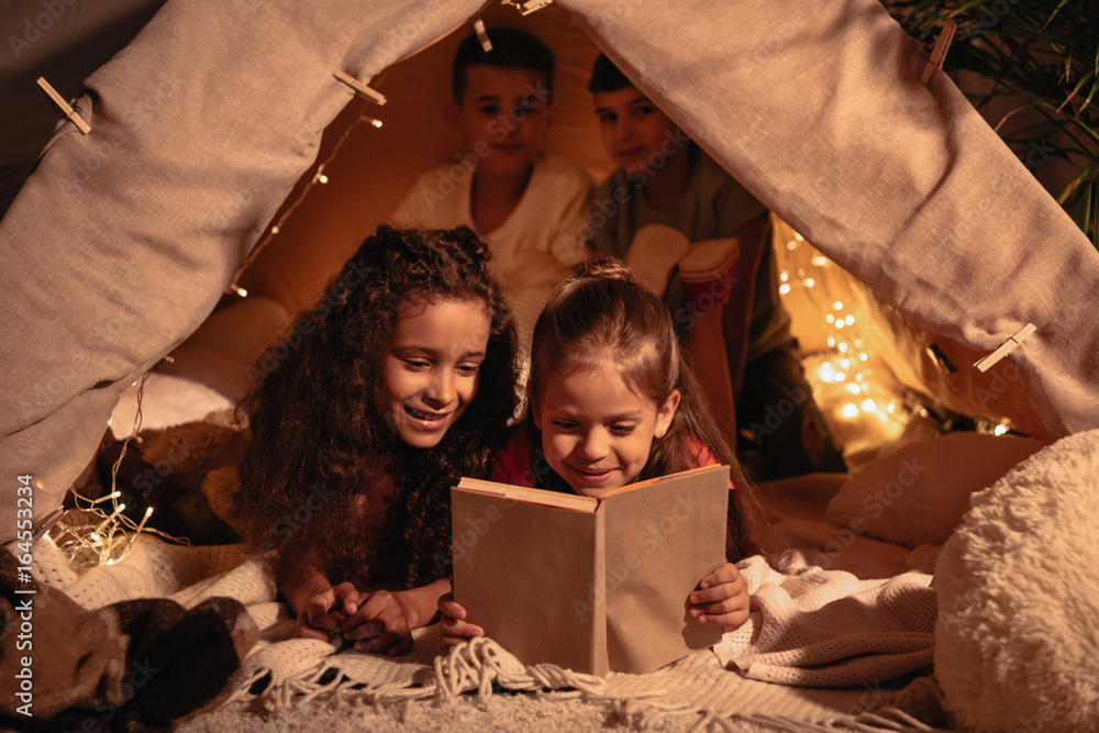 adorable girls reading book together while resting in handmade tent at home