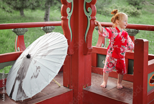 Small toddler redhead girl in traditional Japan National Costume yukata with oil-paper umbrella Wagasa or Bangasa on the background of Asian alcove. multicultural society concept photo