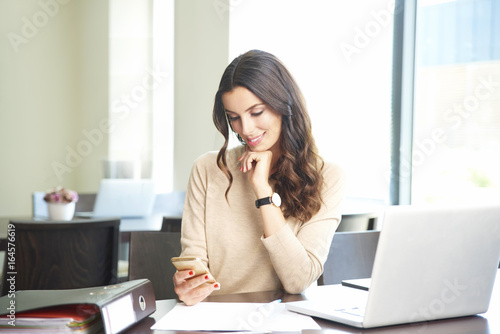 Working hard in the office. Shot of a young financial consultant doing some paperwork and text messaging while sitting at office desk. © gzorgz