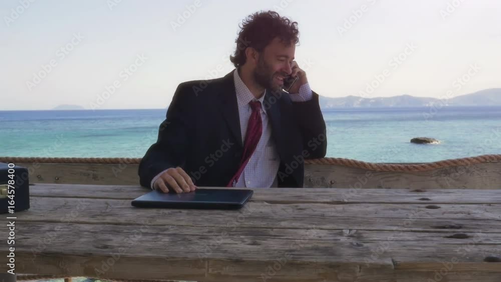 Man in suit talking on his smartphone on a balcony on an island in front of the ocean with laptop Man in suit talking on his smartphone on a balcony on an island in front of the ocean with laptop