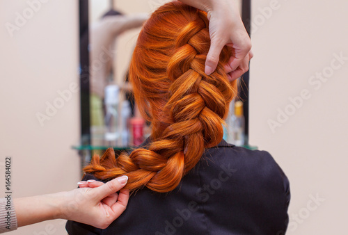 Beautiful, red-haired girl with long hair, hairdresser weaves a French braid, in a beauty salon. Professional hair care and creating hairstyles. photo