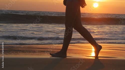 Legs of young woman going along ocean beach during sunrise. Female feet walking barefoot on sea shore at sunset. Girl stepping in shallow water at shoreline. Summer vacation concept. Close up