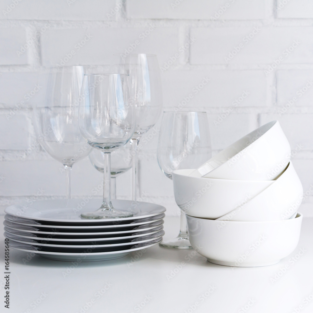 White dishware stacked on a wooden table against white background with transparent wineglasses. Copy space. White dishware stacked on a wooden table against white background with transparent wineglasses. Copy space.