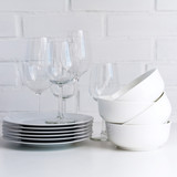 White dishware stacked on a wooden table against white background with transparent wineglasses. Copy space.
