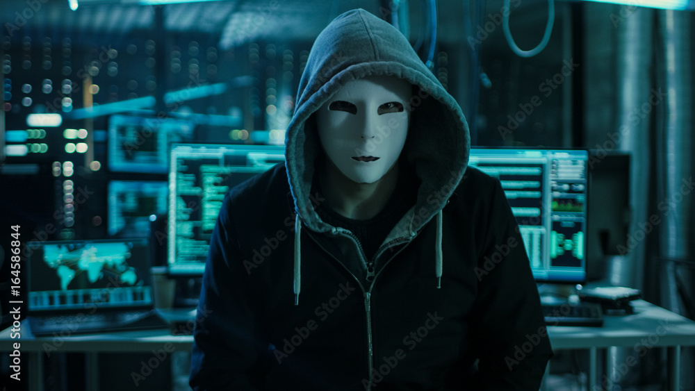 Dangerous Internationally Wanted Hacker with Covered Face Looking into the Camera. In the Background His Operating Room with Multiple Displays and Cables.