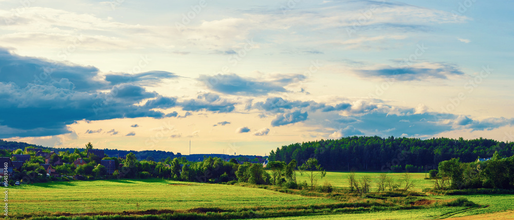 Sunset in the countryside. Rural landscape. Evening sunset sky with clouds, field, trees and farmhouses. Panoramic shot.