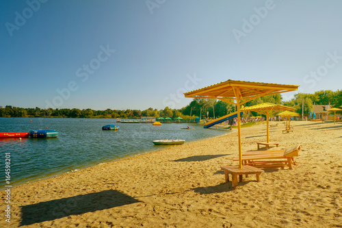 Fototapeta Naklejka Na Ścianę i Meble -  Summer sunny day. Beach, Wooden umbrella, sun loungers on the shore of a lake on the blue sky background.