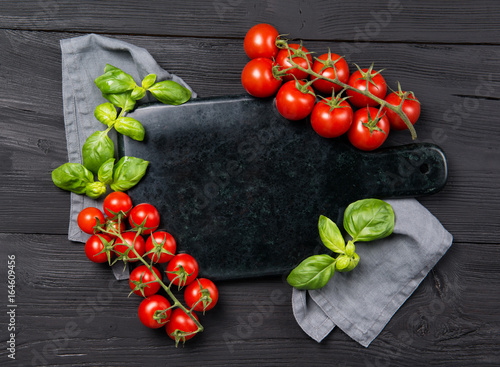 Empty marble cutting board with frame made of ripe red cherry tomatoes and basil leaves, black wooden background, top view with copy space
