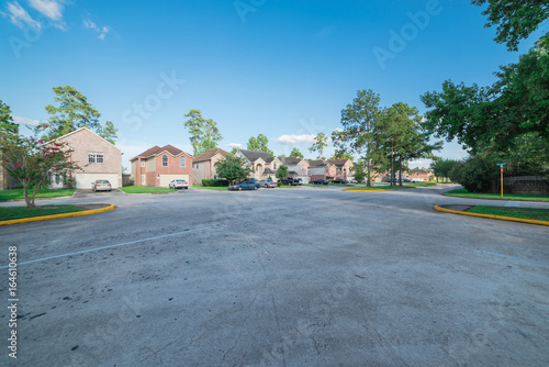 Suburban residential area, row of modern townhomes in Humble, Texas, US. Red brick houses surrounded with tall pine trees, cloud blue sky. Wide view from street intersection of multi-story townhouses.