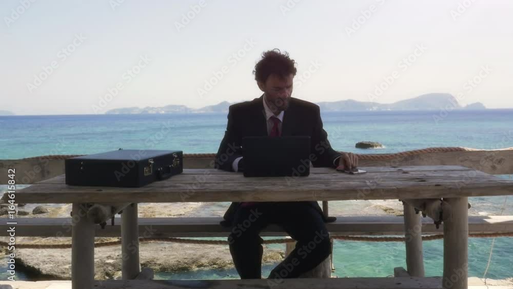 Man in suit talking on his smartphone on a balcony on an island in front of the ocean with laptop Man in suit talking on his smartphone on a balcony on an island in front of the ocean with laptop
