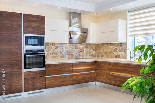 Modern home interior. Expensive, elegant kitchen furniture design. Cabinet facades are made from natural walnut veneer. Patterned wall tiles. Wall-mounted stainless steel cooker hood. Wood cupboard.