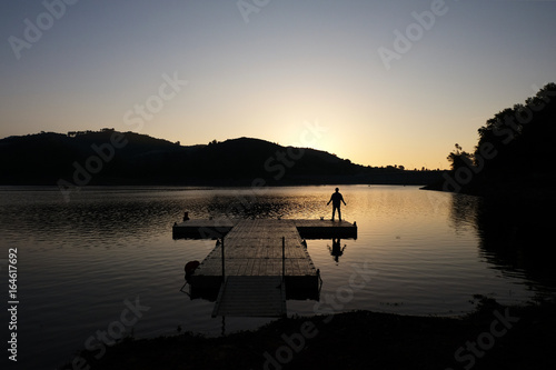 Hombre esperando la salida del sol en un pequeño pantalán o pasarela de madera en un pantano de Cataluña