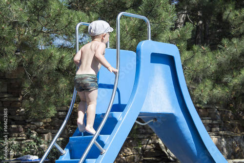 Fototapeta Naklejka Na Ścianę i Meble -  Happy little boy climbing on slide in the pool. children playground near the swimming pool. summer outdoors.